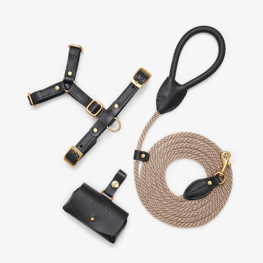 Minimalist flatlay of the black leather dog walking set by CacheMio & CHOUCHOÚ, featuring a harness, rope leash, and poop bag holder.