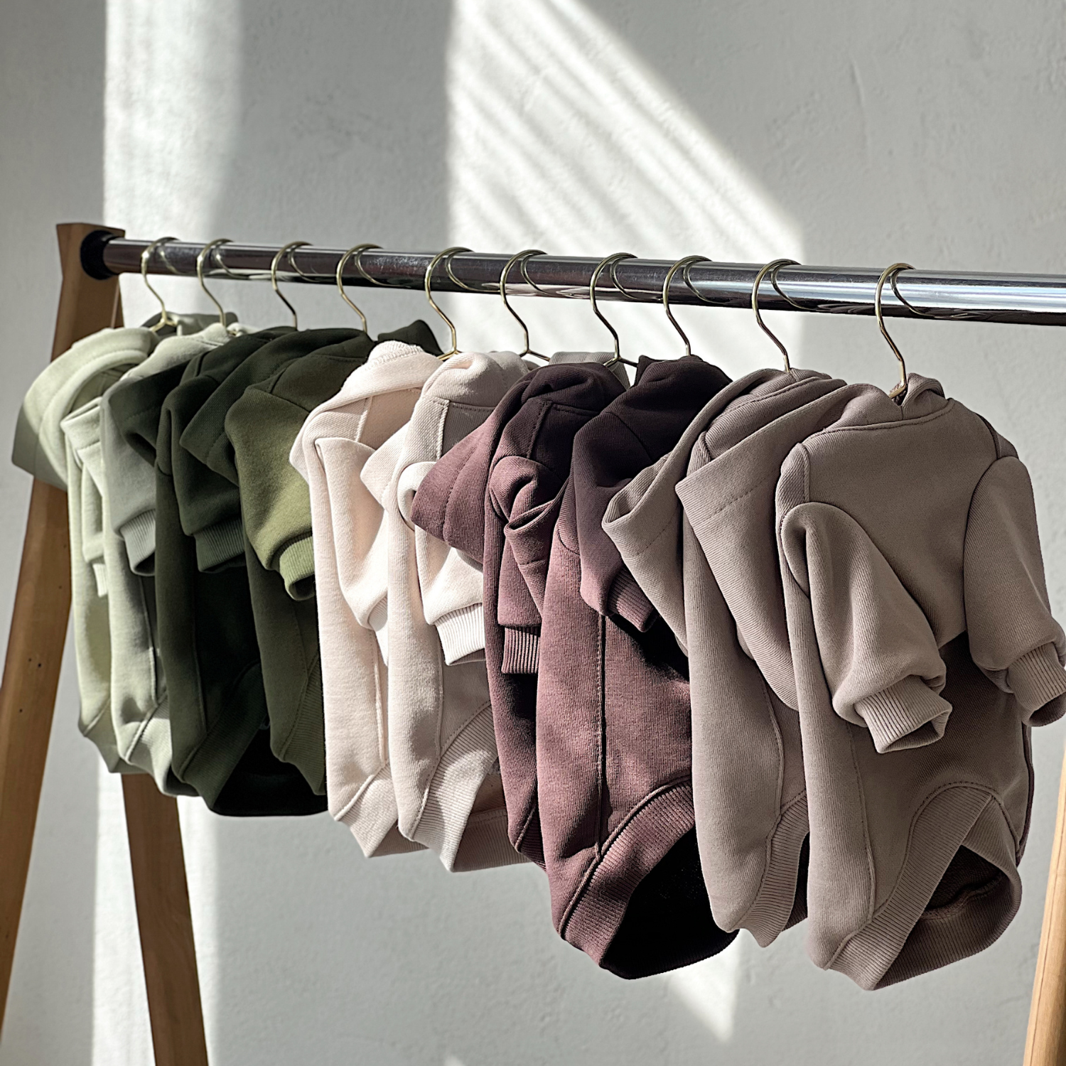 An aesthetic wooden garment rack gracefully displays an entire spectrum of earth-toned dog hoodies, perfectly curated from soft sage green to deep espresso brown.