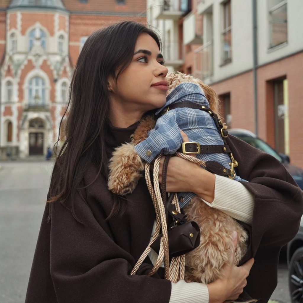 A woman in a brown coat affectionately hugs her brown maltipoo, who is wearing a blue plaid dog shirt and a brown leather harness.
