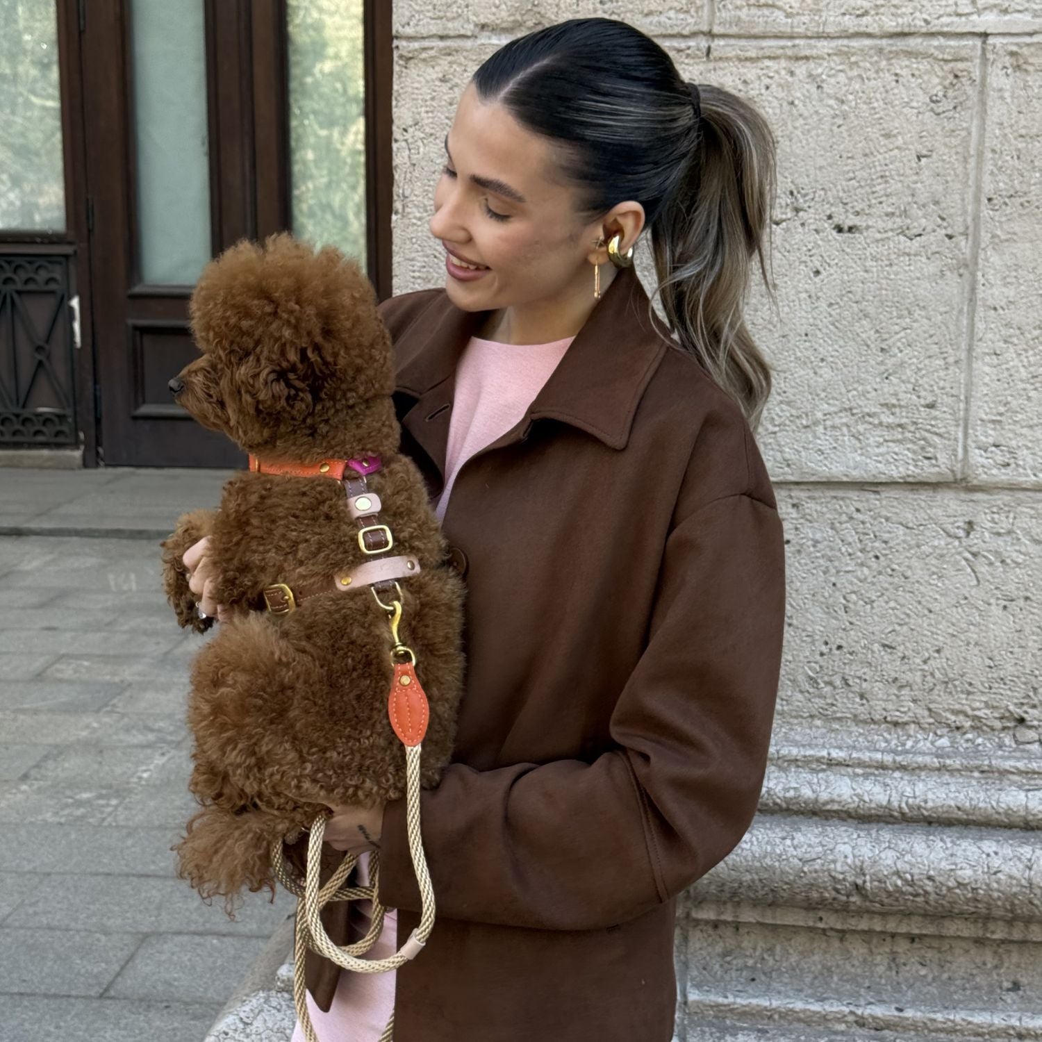 Smiling owner in a brown jacket affectionately holds her maltipoo, showcasing the vivid tangerine and pink accents of the avant-garde pet vest.