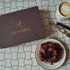 Closed brown magnetic gift box featuring the CHOUCHOÚ logo, styled aesthetically on a cafe table with coffee and a pastry.