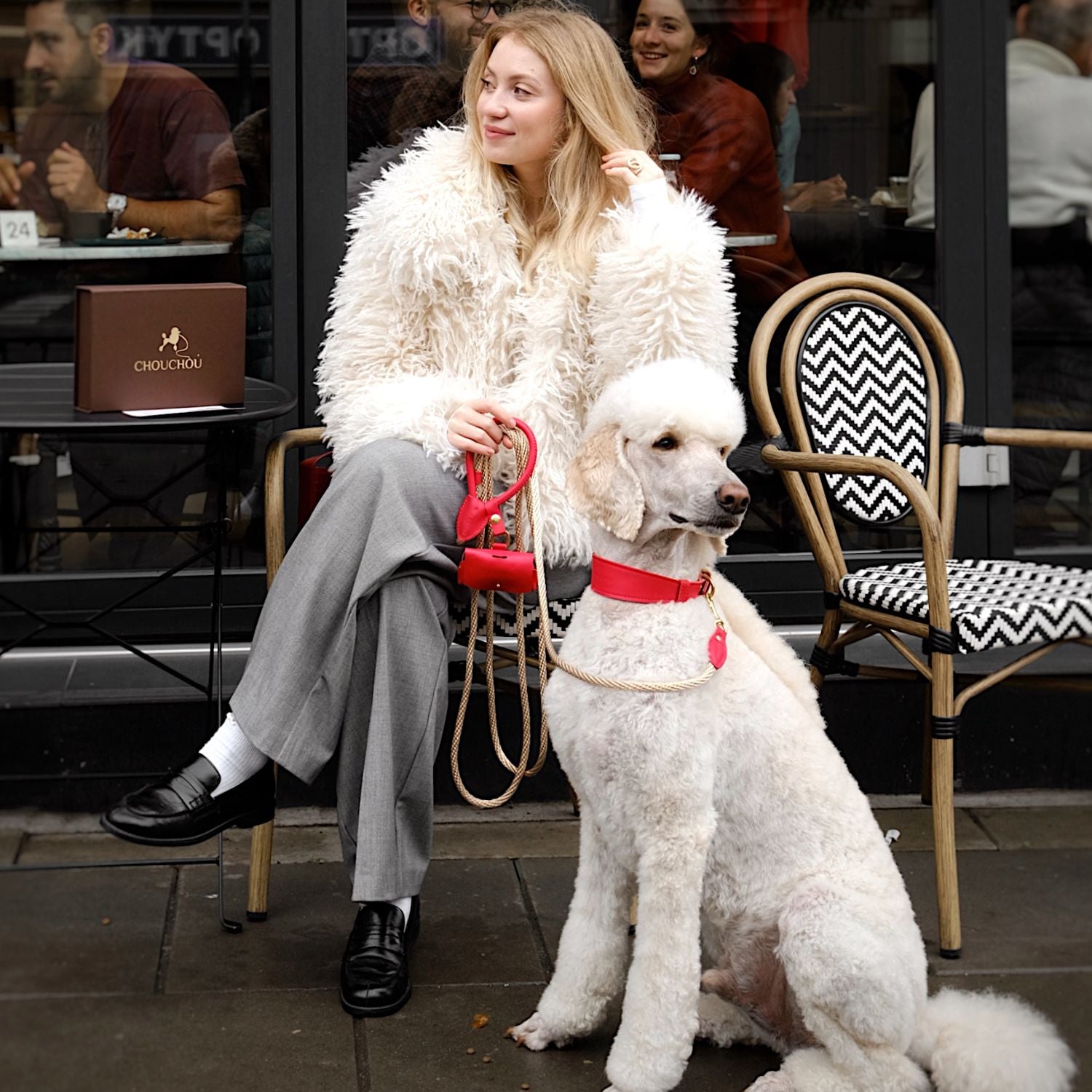 Elegant standard white poodle wearing premium red leather dog accessories by CacheMio & CHOUCHOÚ, sitting next to its owner at a European city cafe.