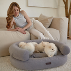 Relaxed domestic harmony shown as a woman reclines on a modular sofa, observing tiny white Pomeranian puppies settling into the spacious silver bed.