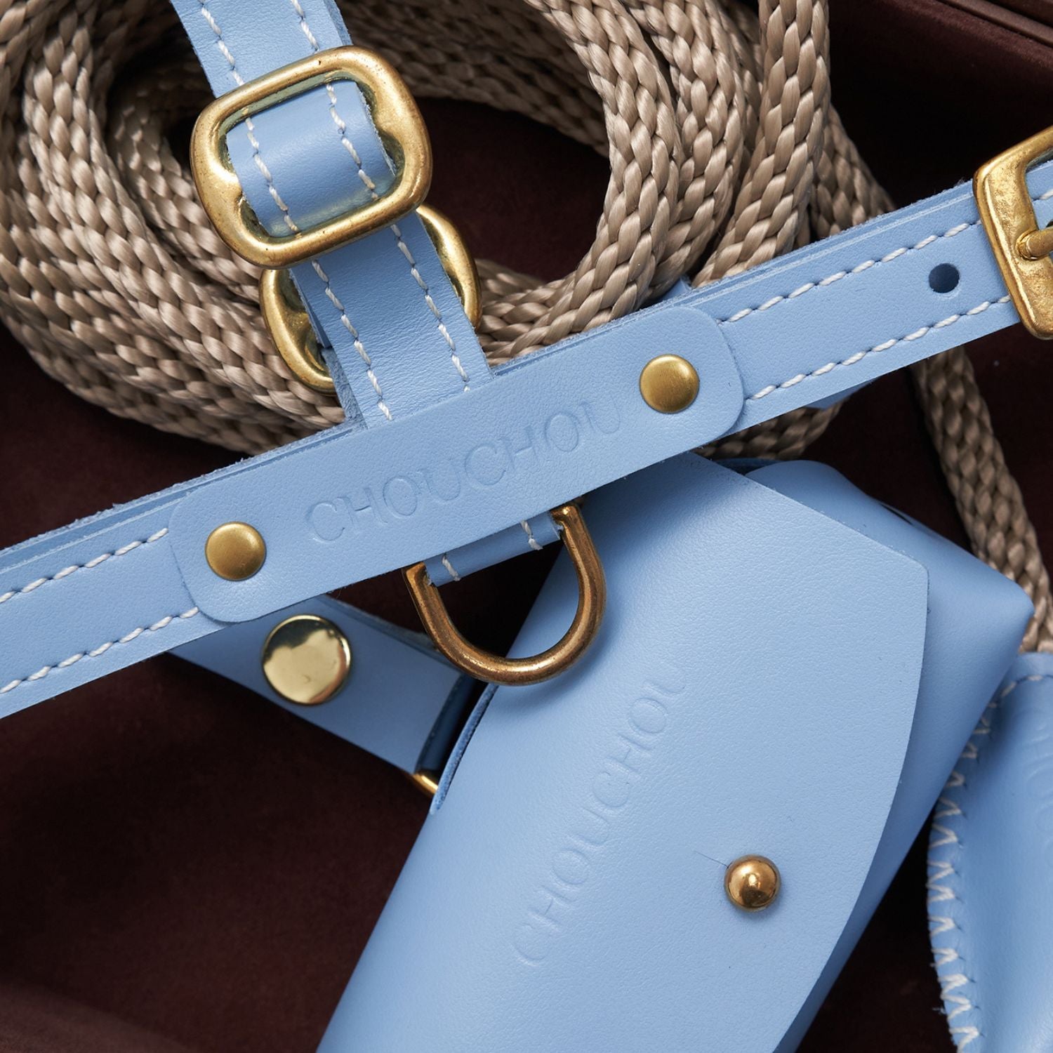 Macro photography of the CHOUCHOÚ light blue leather dog harness, showcasing the embossed logo, meticulous stitching, and solid brass D-ring.