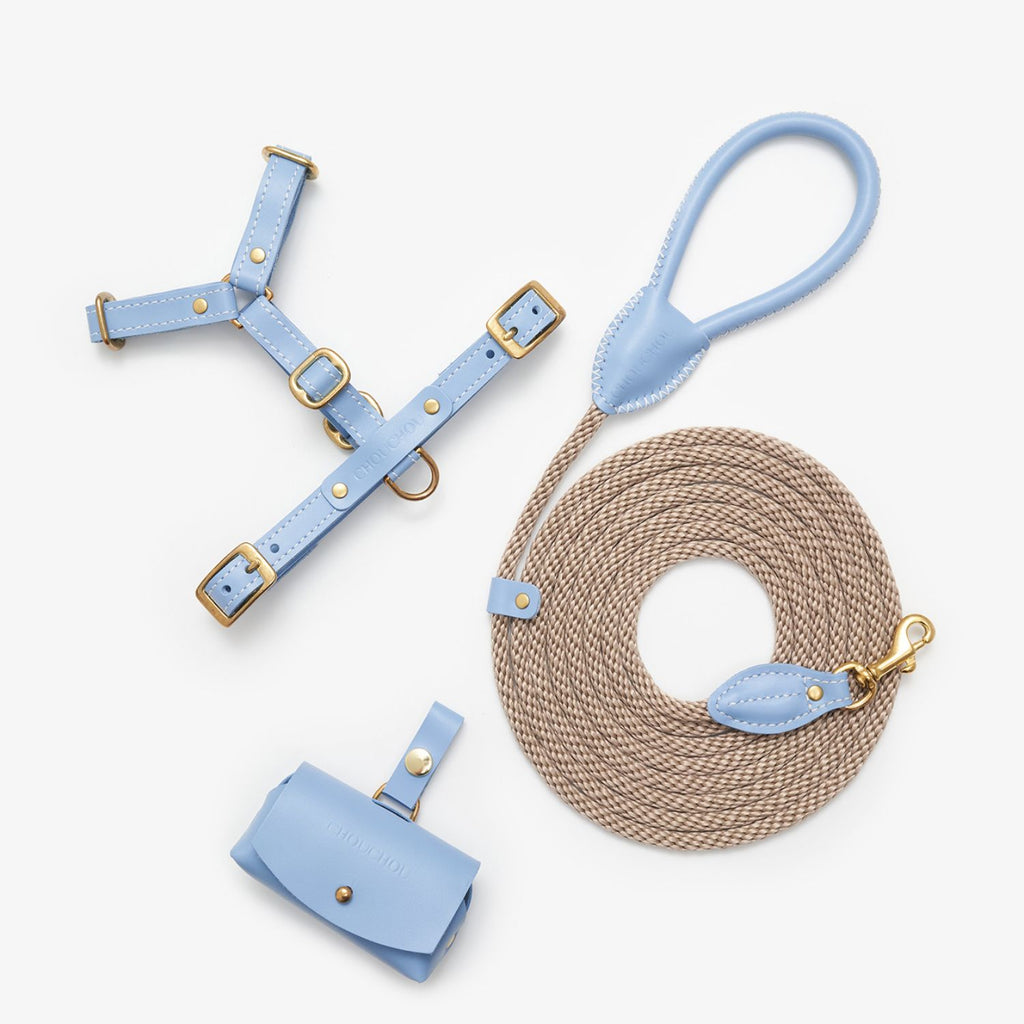 Minimalist flatlay of the CHOUCHOÚ light blue dog walking set, featuring a leather harness, rope leash, and poop bag dispenser.