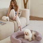Pet owner relaxing on a beige sofa with her dog, while another white Bichon Frise enjoys the supportive pink Marshmallow bed by CHOUCHOÚ.